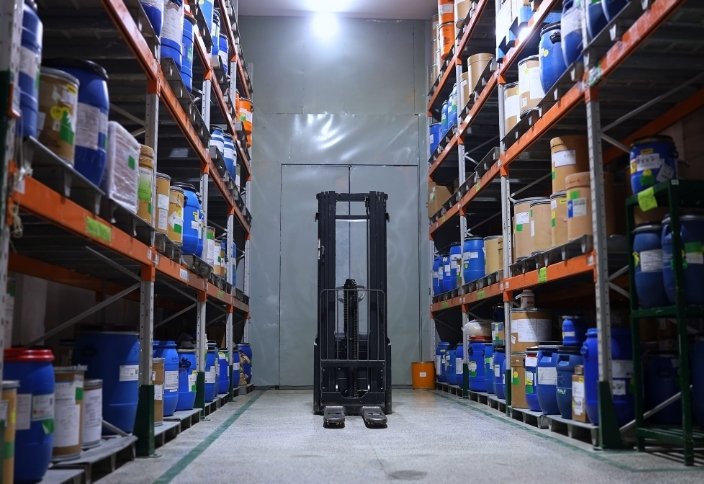 Armazém de produtos químicos industriais com recipientes organizados em estantes e um empilhador no corredor.